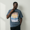 male model against a wall wearing a blue t-shirt with a sunset graphic design