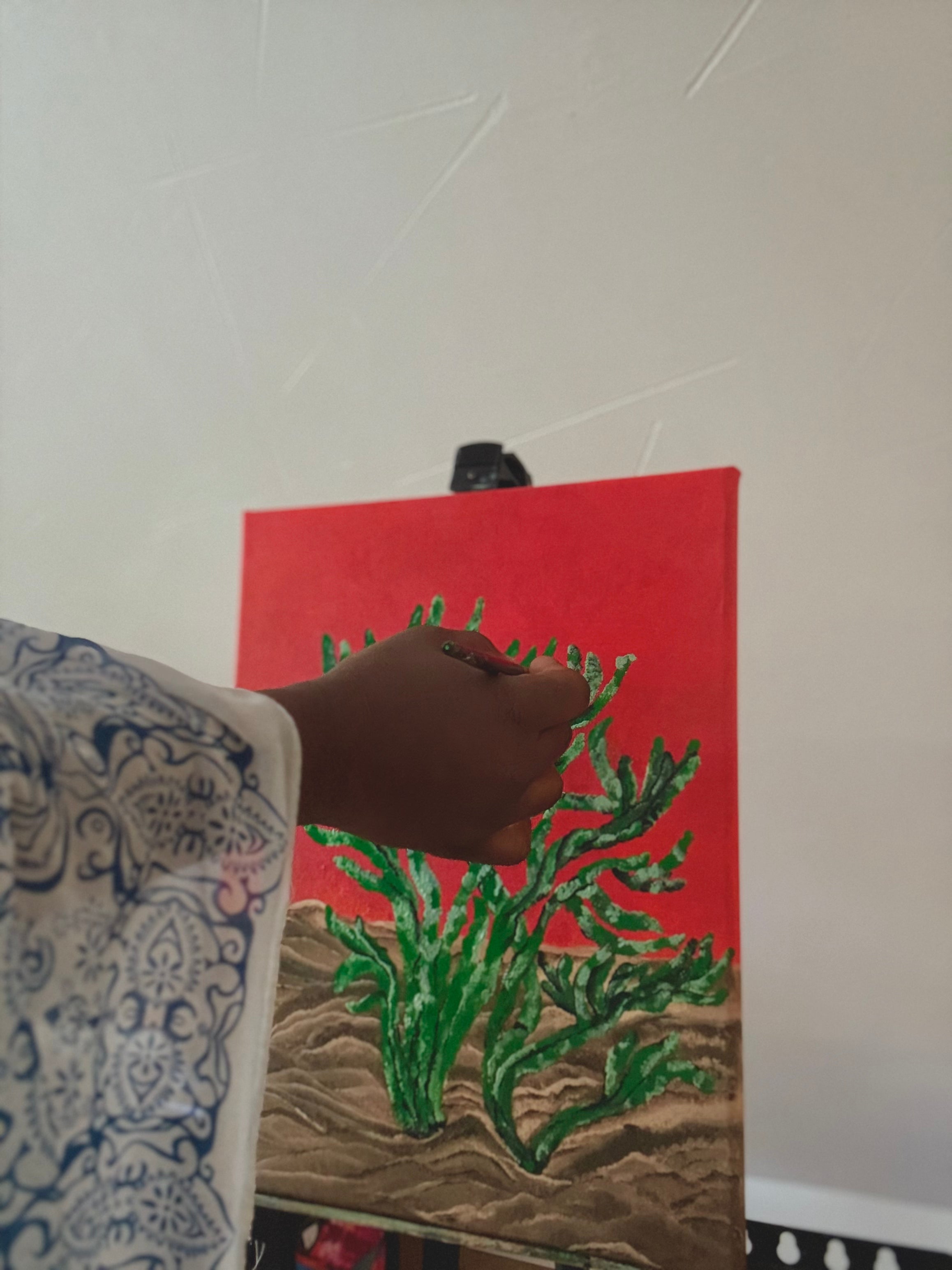 A hand holding a paintbrush painting on a canvas, which features a bright red sky and green cactus-like plants on a sandy brown ground.