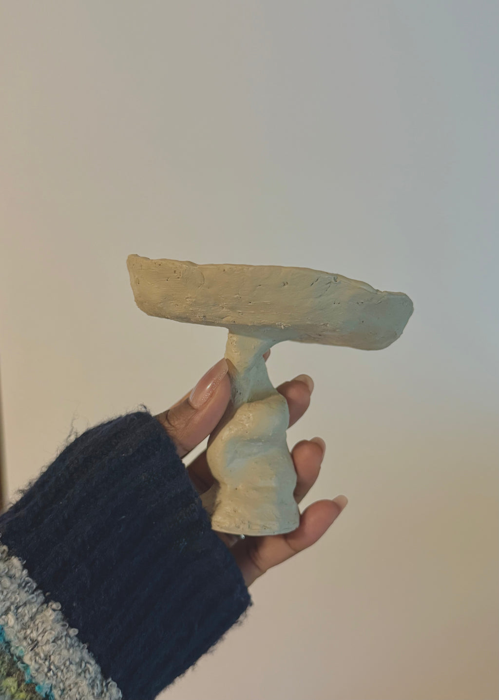 Handcrafted-style sculptural bowl with a twisted pedestal base, held in a person's hand wearing a dark blue knit sleeve against a plain off-white wall.